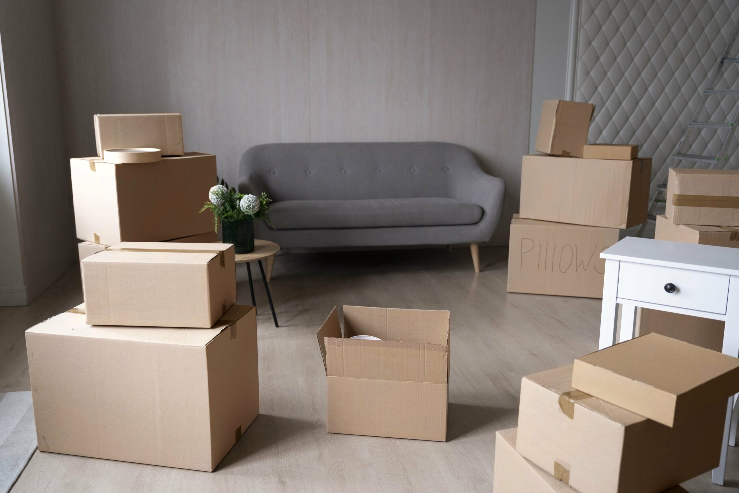 Salon lumineux avec plusieurs boîtes en carton prêtes à emballer, quelques colis ouverts et une petite table avec fleurs, prête pour un déménagement. Inclure le conseil: conseil preparation demenagement.